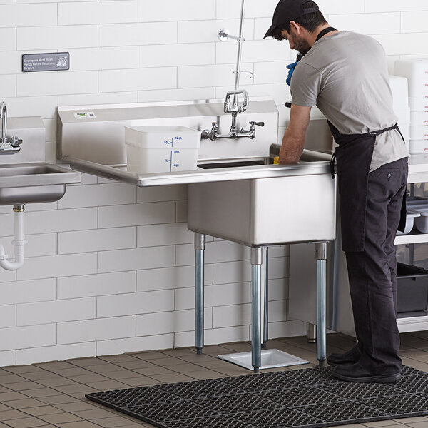 A man in an apron washing dishes in a Regency stainless steel sink with a left drainboard in a professional kitchen.