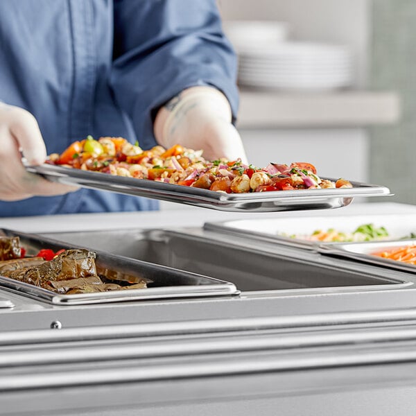 A person in a chef's uniform holding a Vollrath stainless steel steam table pan of food.