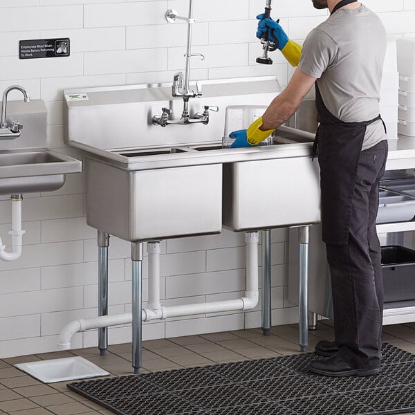 A man in a black shirt and blue jeans standing in a professional kitchen using a Regency 2 compartment stainless steel sink.