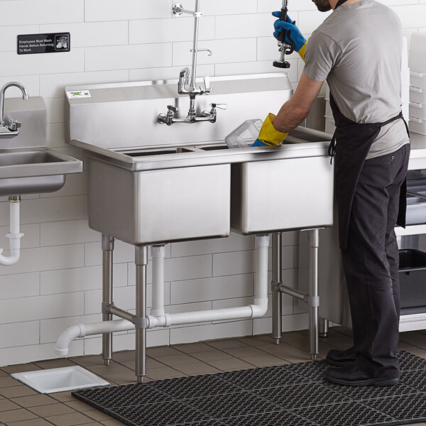 A man in a kitchen wearing an apron and gloves washing a Regency two compartment sink.