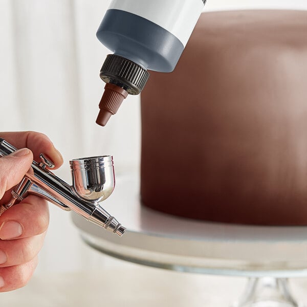 A person using a silver Chefmaster airbrush to spray brown color onto a cake.