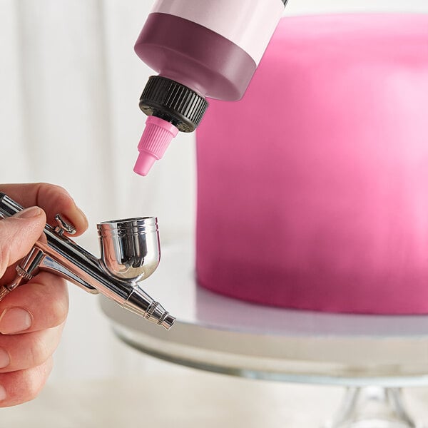 A finger holding a Chefmaster Neon Brite Pink airbrush color bottle over a cake.