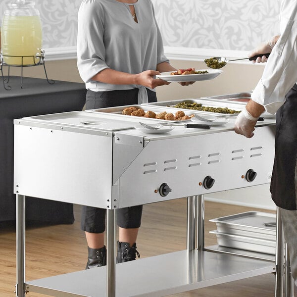 A woman serving food to a man from an Avantco mobile steam table on a table in an outdoor catering setup.