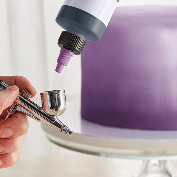 A person using a silver airbrush to spray purple color over a cake.