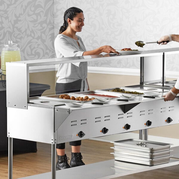 A woman serving food at a hotel buffet using an Avantco open well steam table with sneeze guard.