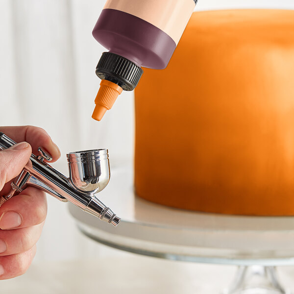 A finger holding a bottle of Chefmaster Neon Brite Orange airbrush color.