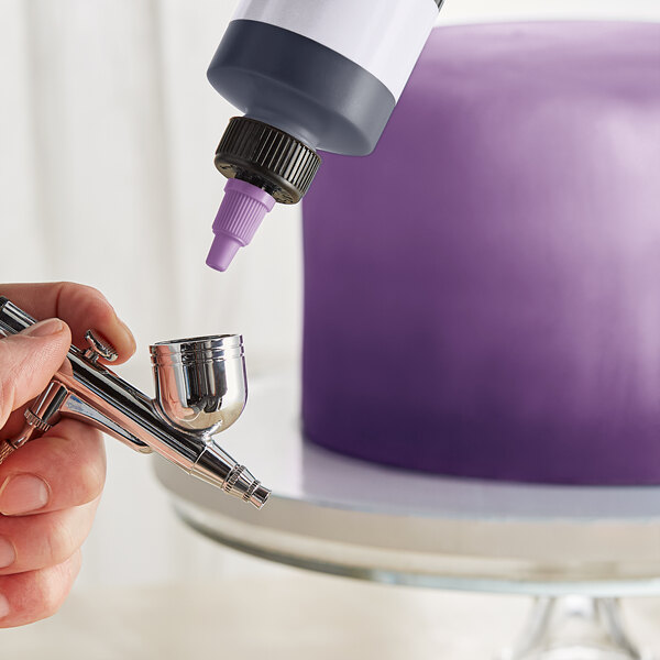 A person using a silver airbrush to spray purple color over a cake.