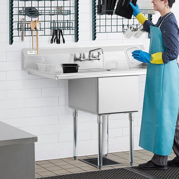 A woman in a blue apron using a Regency stainless steel one compartment sink with drainboards.