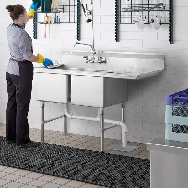 A woman wearing a black apron and yellow gloves washing dishes in a Regency stainless steel 2 compartment sink with a right drainboard.