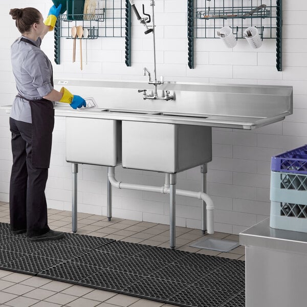 A woman in a professional kitchen cleaning a Regency stainless steel 2 compartment sink.
