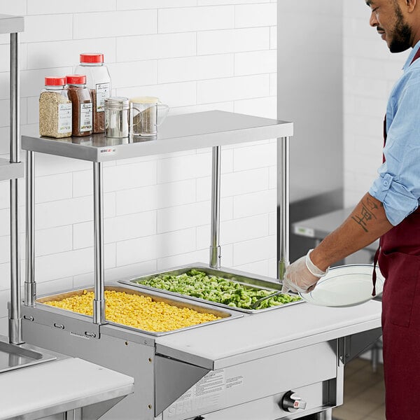 A man in a red apron standing in a kitchen in front of a ServIt single overshelf above a steam table.