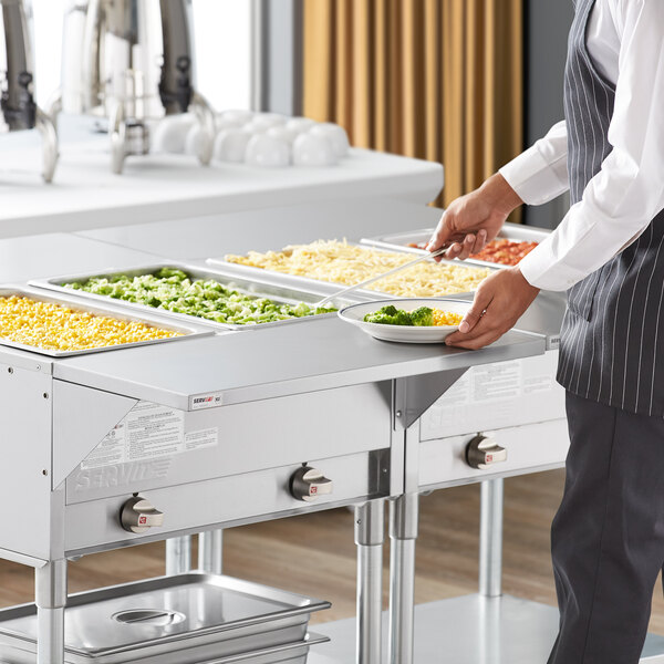 A man in a chef's uniform serving food using a ServIt solid metal tray on a steam table.