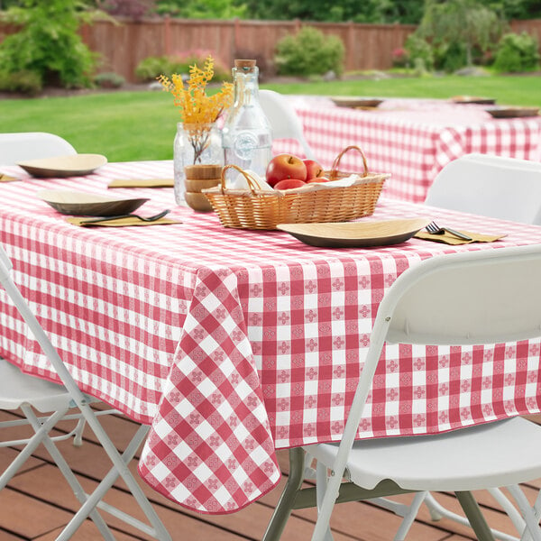 A picnic table with a red and white checkered vinyl table cover, white plates, and a basket of fruit.