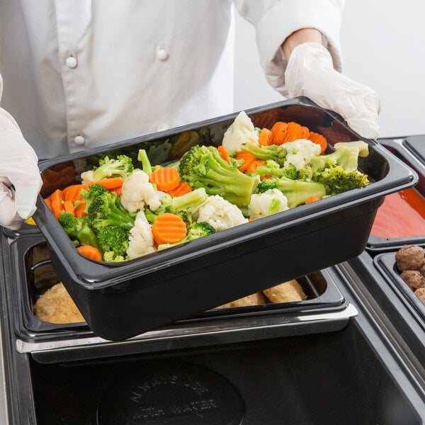 A person in white gloves holding a Cambro black plastic food pan filled with vegetables.