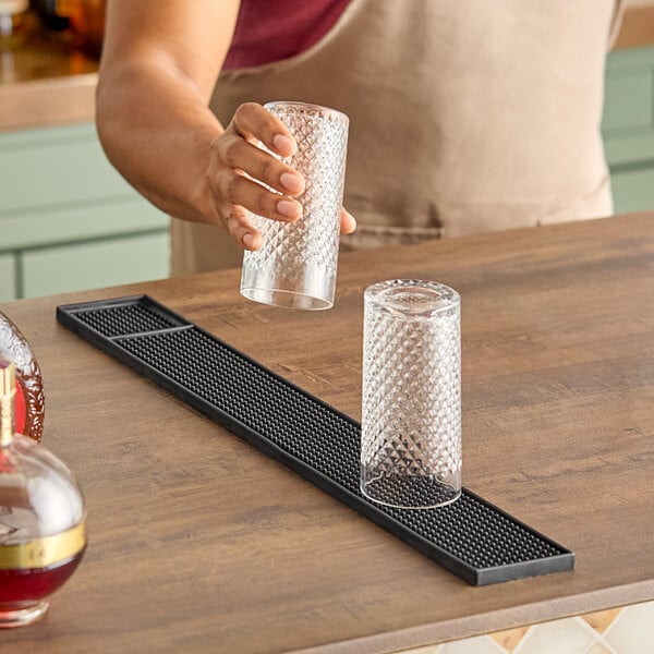 A hand holding a glass over a Vollrath black bar mat.