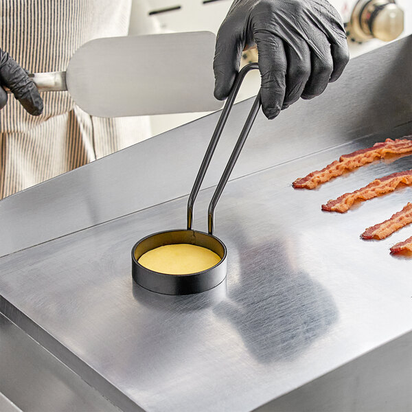 A person in black gloves using a Vigor 3" egg ring to cook bacon on a grill.