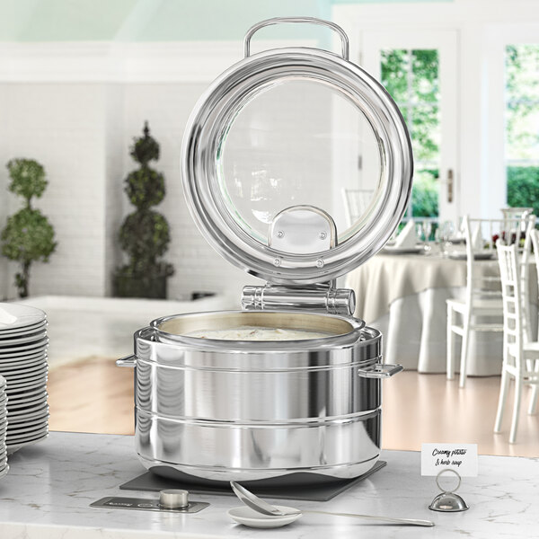 An Acopa Manchester stainless steel chafer with a glass top on a table with plates and chairs.