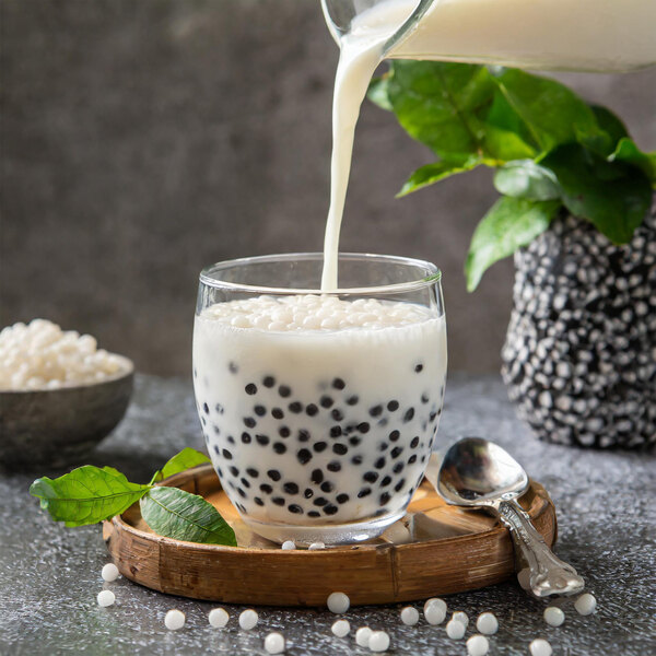 A glass of milk with black tapioca balls being poured into a bowl of white liquid.