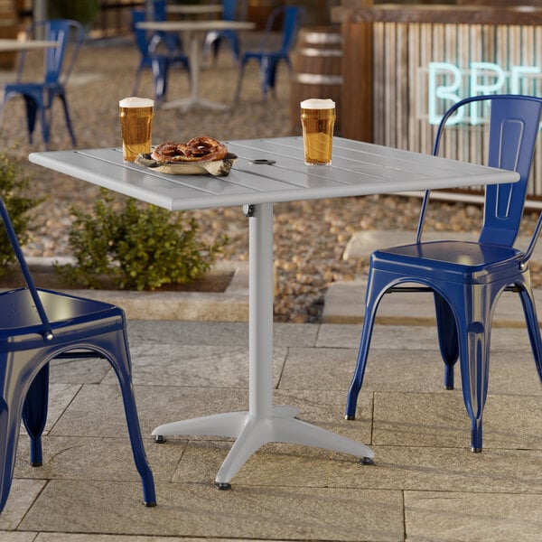 A square silver powder-coated aluminum outdoor dining table with an umbrella hole, surrounded by blue metal chairs.