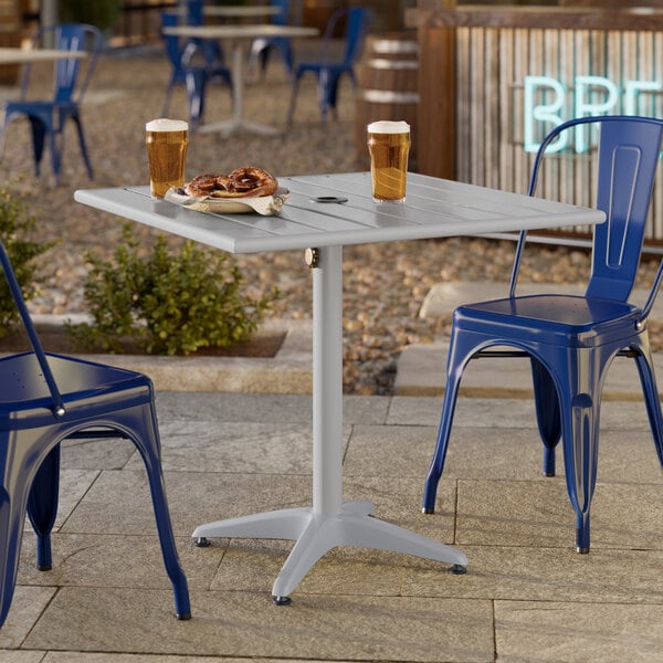 A square silver powder-coated aluminum outdoor dining table with an umbrella hole, shown with two blue metal chairs.