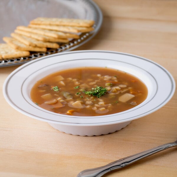 A white plastic soup bowl with silver bands filled with soup and crackers on a table.