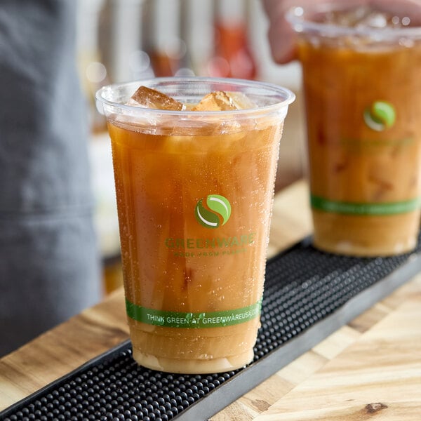 A clear 20 oz. compostable plastic cold cup filled with iced coffee, featuring Greenware branding.