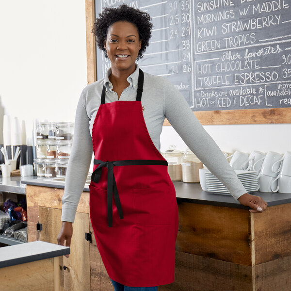 A smiling woman wearing a Choice red poly-cotton bib apron with black webbing accents.