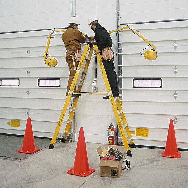 A man in brown overalls and a tool belt holding a yellow Bauer 2-way step ladder with another man on it.