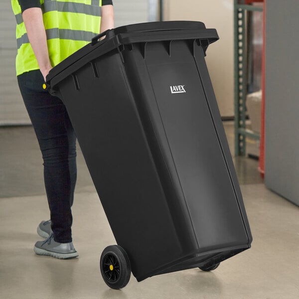 A person in a yellow vest pushing a large black Lavex rectangular trash can.