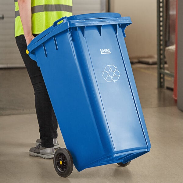 A person in a yellow vest pushing a Lavex blue wheeled rectangular recycle bin.