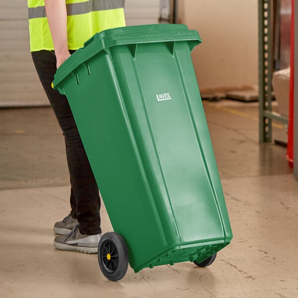 A woman in a yellow vest pushing a Lavex green wheeled rectangular trash can.