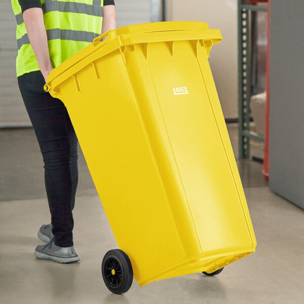 A woman in a yellow shirt pushing a Lavex yellow wheeled rectangular trash can.