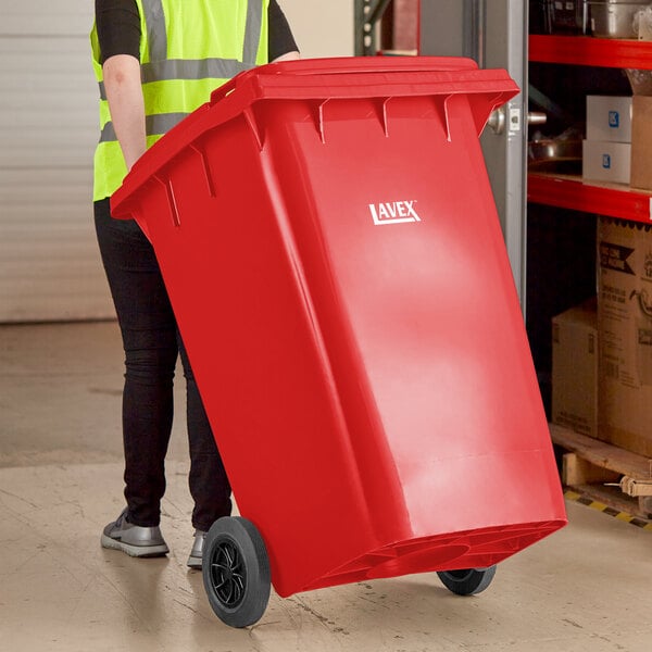 A woman in a yellow vest pushing a Lavex red rectangular trash can with wheels.