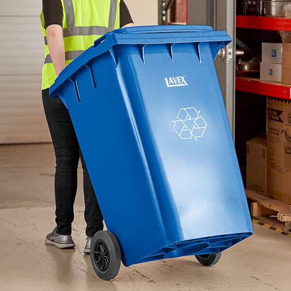 A woman in a yellow vest pushing a Lavex blue wheeled rectangular recycle bin.