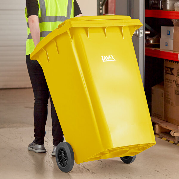 A woman in a yellow vest pushing a Lavex yellow wheeled rectangular trash can.