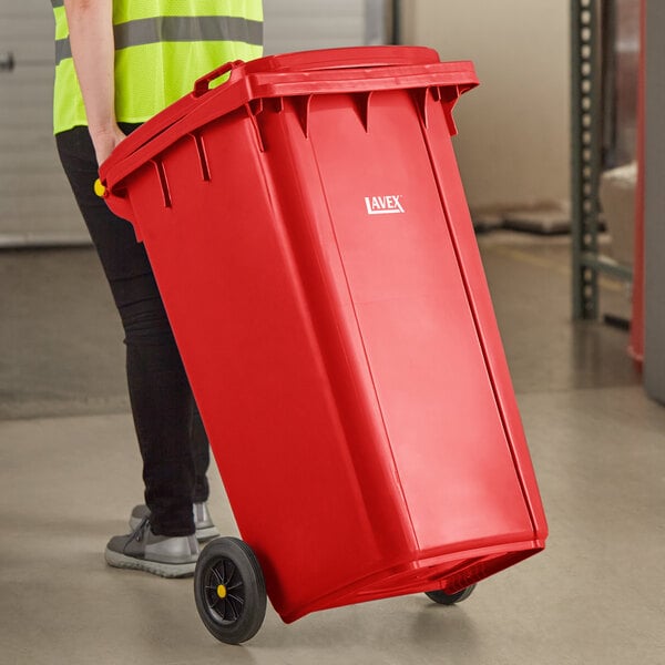 A woman in a yellow vest pushing a red Lavex rectangular trash can with wheels.