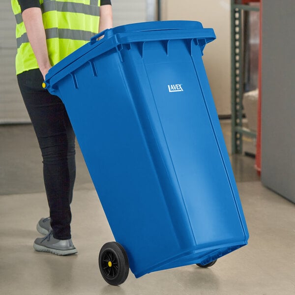 A person in a yellow vest pushing a Lavex blue rectangular trash can with wheels.