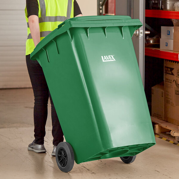 A woman in a green vest pushing a Lavex green wheeled rectangular trash can.