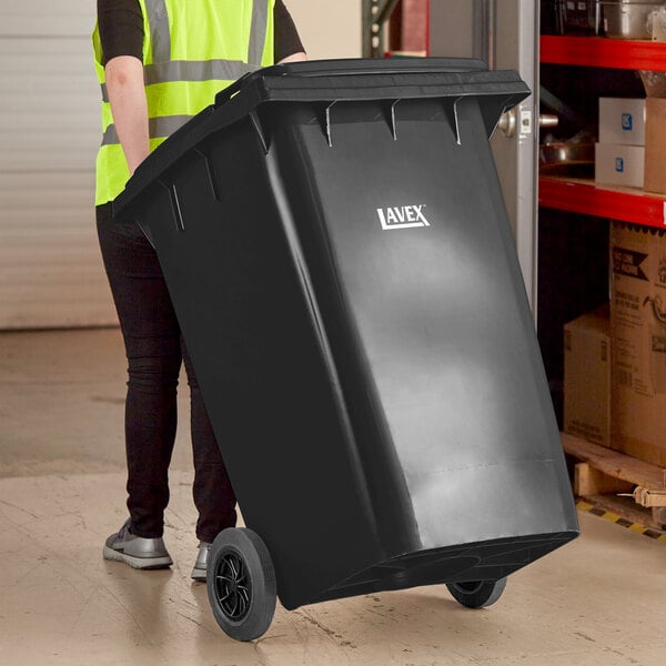 A woman in a yellow vest pushing a large black Lavex wheeled trash can.