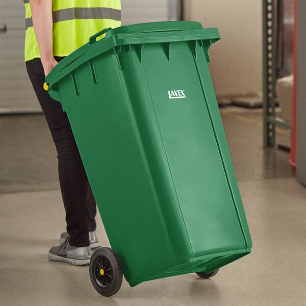 A woman pushing a Lavex green wheeled rectangular trash can.