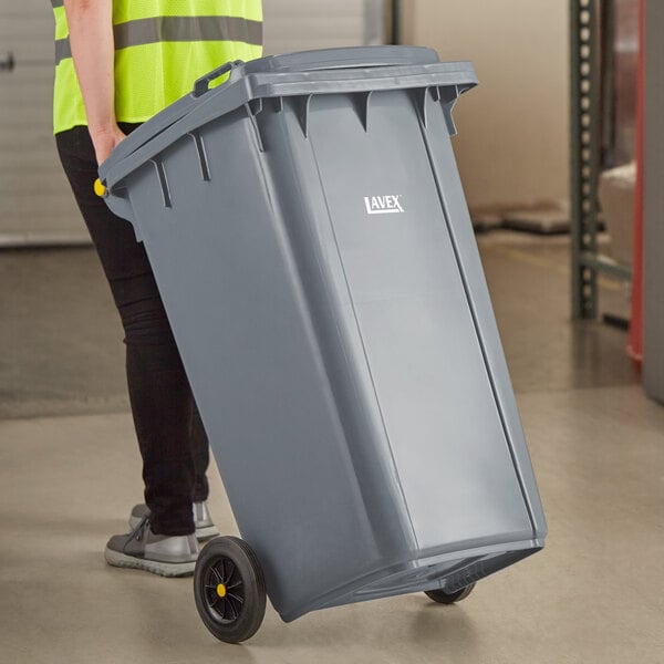 A woman in a yellow vest pushing a Lavex gray wheeled rectangular trash can.