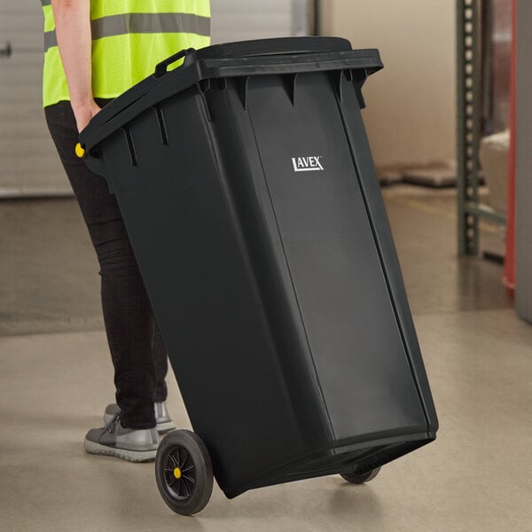 A woman in a safety vest pushing a Lavex black wheeled rectangular trash can.