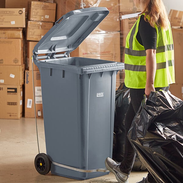 A woman in a yellow vest using a Lavex step-on attachment on a trash can.