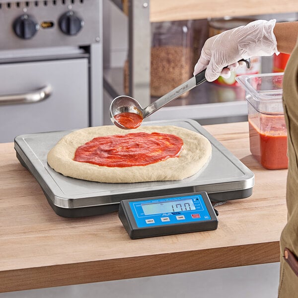 A person using an AvaWeigh digital scale to measure pizza dough next to a container of tomato sauce.
