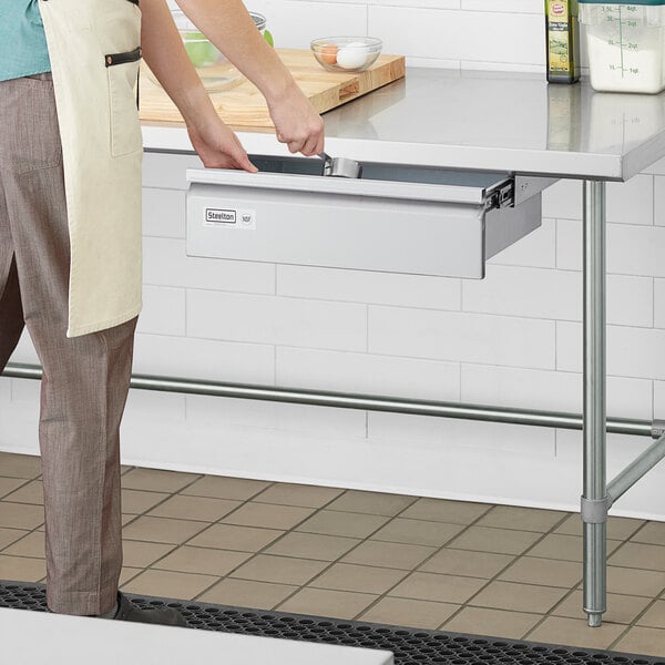 A person wearing a white apron opens a Steelton stainless steel drawer on a kitchen counter.