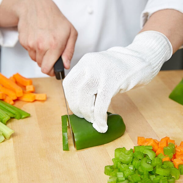 A person wearing Mercer Culinary white cut-resistant gloves cutting vegetables on a cutting board.