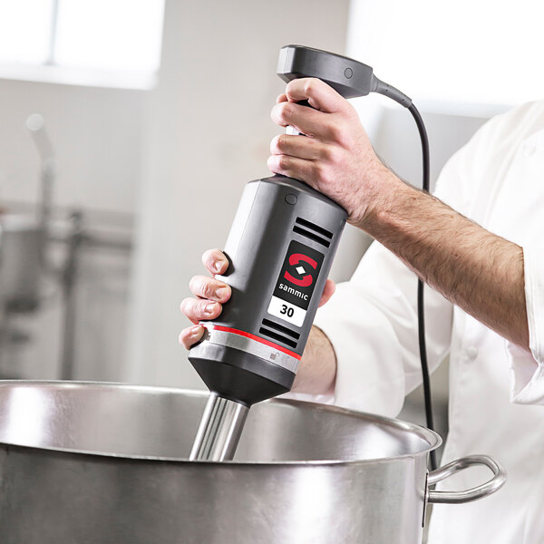 A person using a Sammic medium-duty hand blender to mix a large pot on a counter in a professional kitchen.