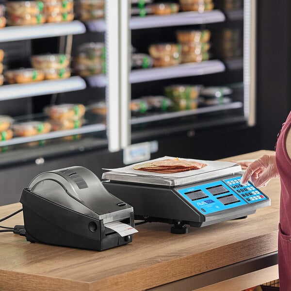 A digital price computing scale with a label printer on a countertop in a grocery or deli setting.