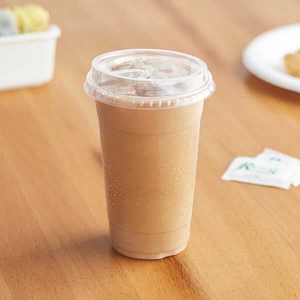 A Choice clear plastic cup with a brown drink inside and a yellow sip-through lid on a table next to cookies.