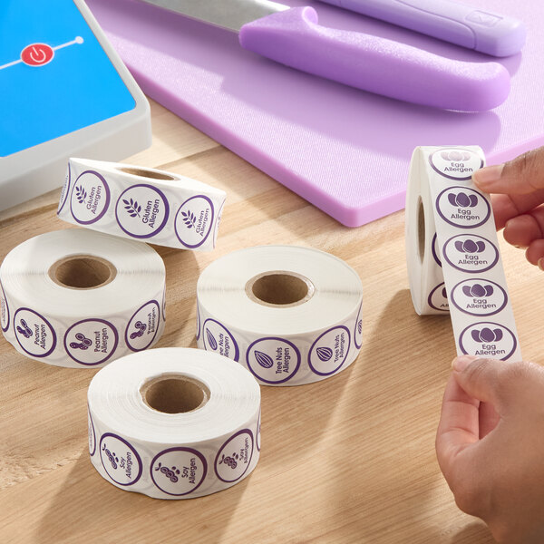 A set of rolls of purple allergen labels, including peanut, gluten, tree nut, and egg allergen warnings, on a wooden surface.